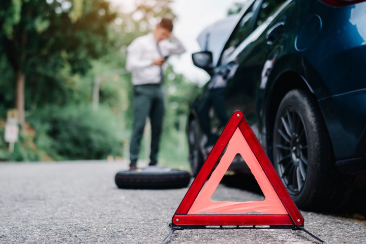 A red reflective warning triangle placed on a roadside in front of a broken-down car, with a blurred person in the background appearing to change a tyre near the vehicle.